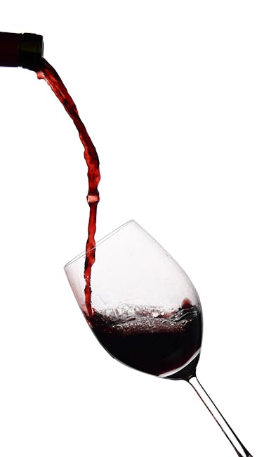 A close-up image of red wine being poured from a bottle into a clear wine glass, positioned on a white surface with a plain white background. The wine appears deep red with a slight sheen, filling the